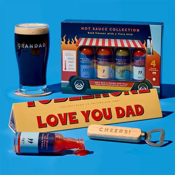 Father’s Day gift arrangement on a blue background featuring a Grandad pint glass, hot sauce gift set, Toblerone bar with Love You Dad message, bottle opener, and spilled sauce bottle